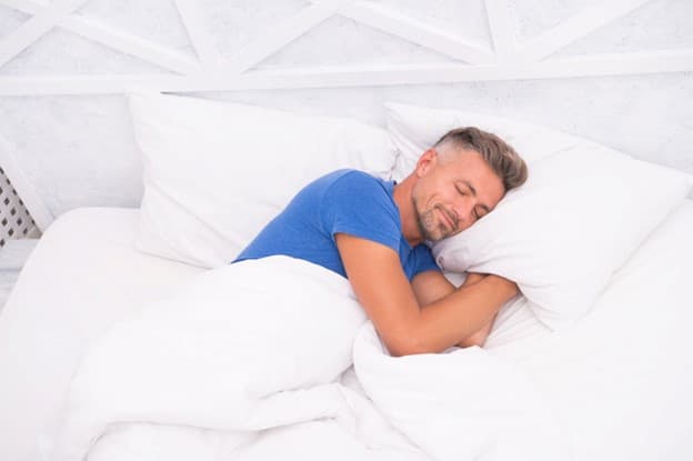 A man with beard and blue t-shirt sleeping in all white bedding